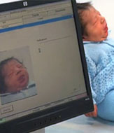 A baby being held next to a computer. The baby's image is reflected on the screen of the computer.