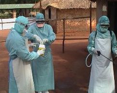 A photograph of three laboratory technicians wearing protective facial masks, gowns, eye visor and gloves while securing a sample container