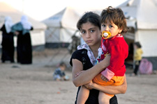 A young girl holds her younger sister against the backdrop of a Syrian refugee camp