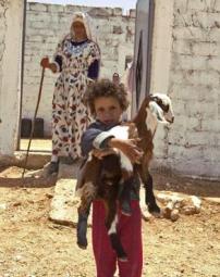 Une petite fille portant une chèvre dans ses bras s'éloigne d'une maison sous l'œil de sa mère debout dans l'encadrement de la porte.