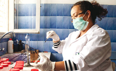 A female health professional in gloves and mask standing at a sink placing some liquid into a test tube 