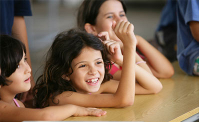 Image of three young girls sitting at a long desk smiling with some other children in the background