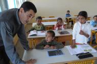 Des enfants sont assis à leur table dans une classe ; au premier plan, le professeur est penché vers les enfants, les deux mains appuyées sur une des tables.