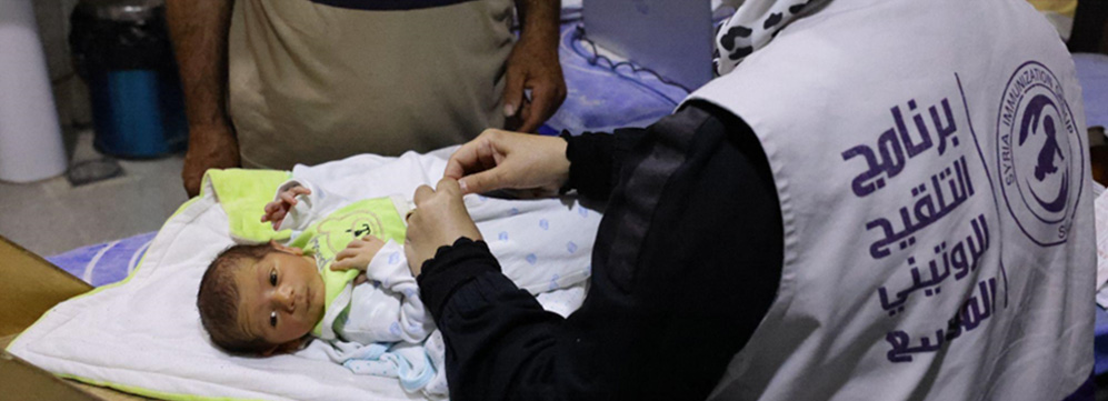 Majed stands beside his newborn baby as a health worker administers a routine vaccine as part of continued efforts to protect children from preventable diseases in conflict-affected areas