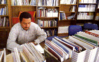 Un homme est assis dans une bibliothèque devant une table chargée de dossiers et de cahiers.