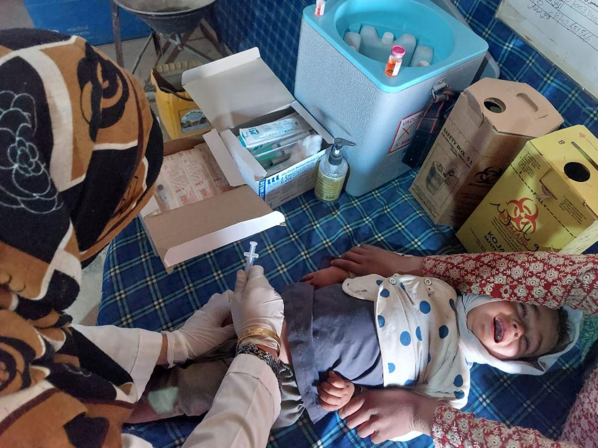 A health worker administers an injection to a child lying on a patterned surface, while another adult gently holds the child. Medical supplies, syringes, and a vaccine cooler are arranged on the table nearby.