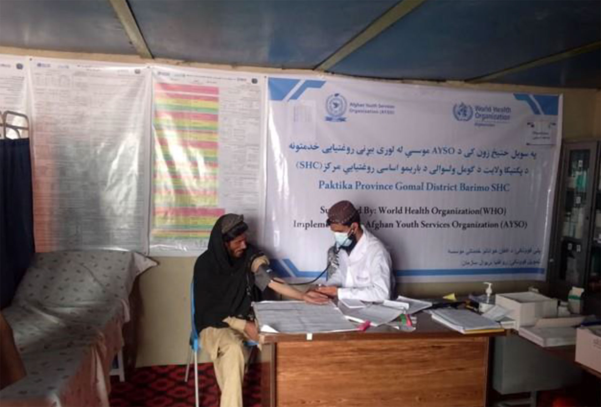 A group of health workers and field staff gather inside a temporary clinic tent, reviewing documents and discussing activities around a central table