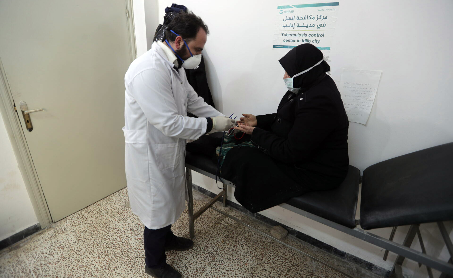 A person in a white coat uses a smartphone to photograph a chest X‑ray displayed on a laptop screen on a desk
