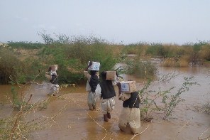 WHO staff and volunteers carrying boxes of medicines and supplies while they cross flooded areas to reach a health facility in Aroma locality, Kassala, East Sudan