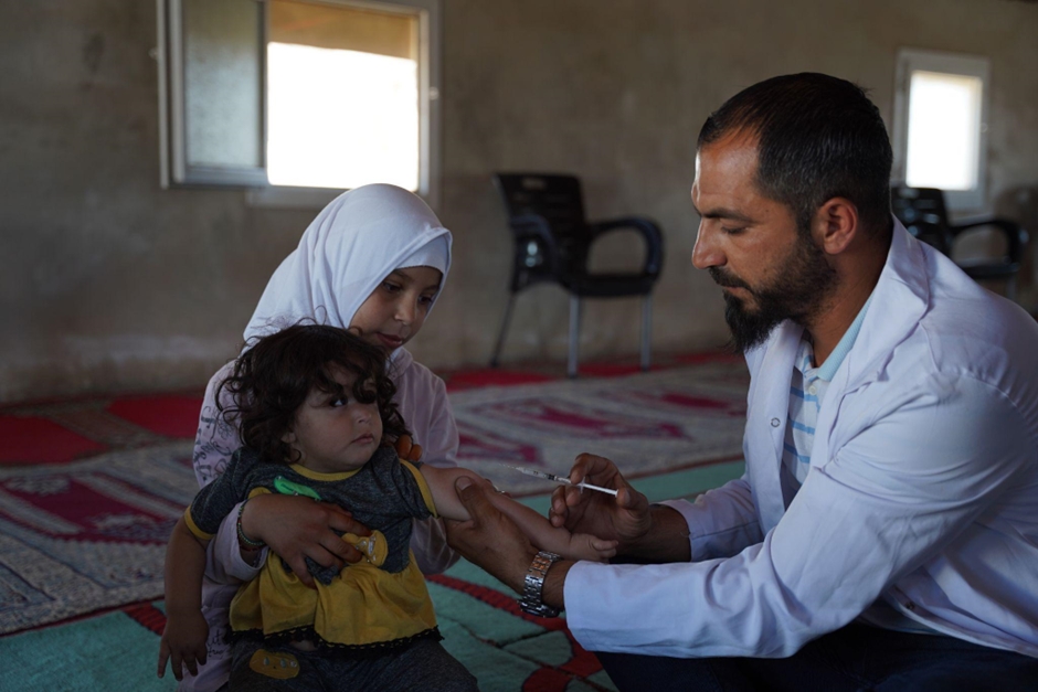   One of Abu Tarek’s daughters holds her younger brother close as Yasser administers a vaccine. Photo credit: WHO 