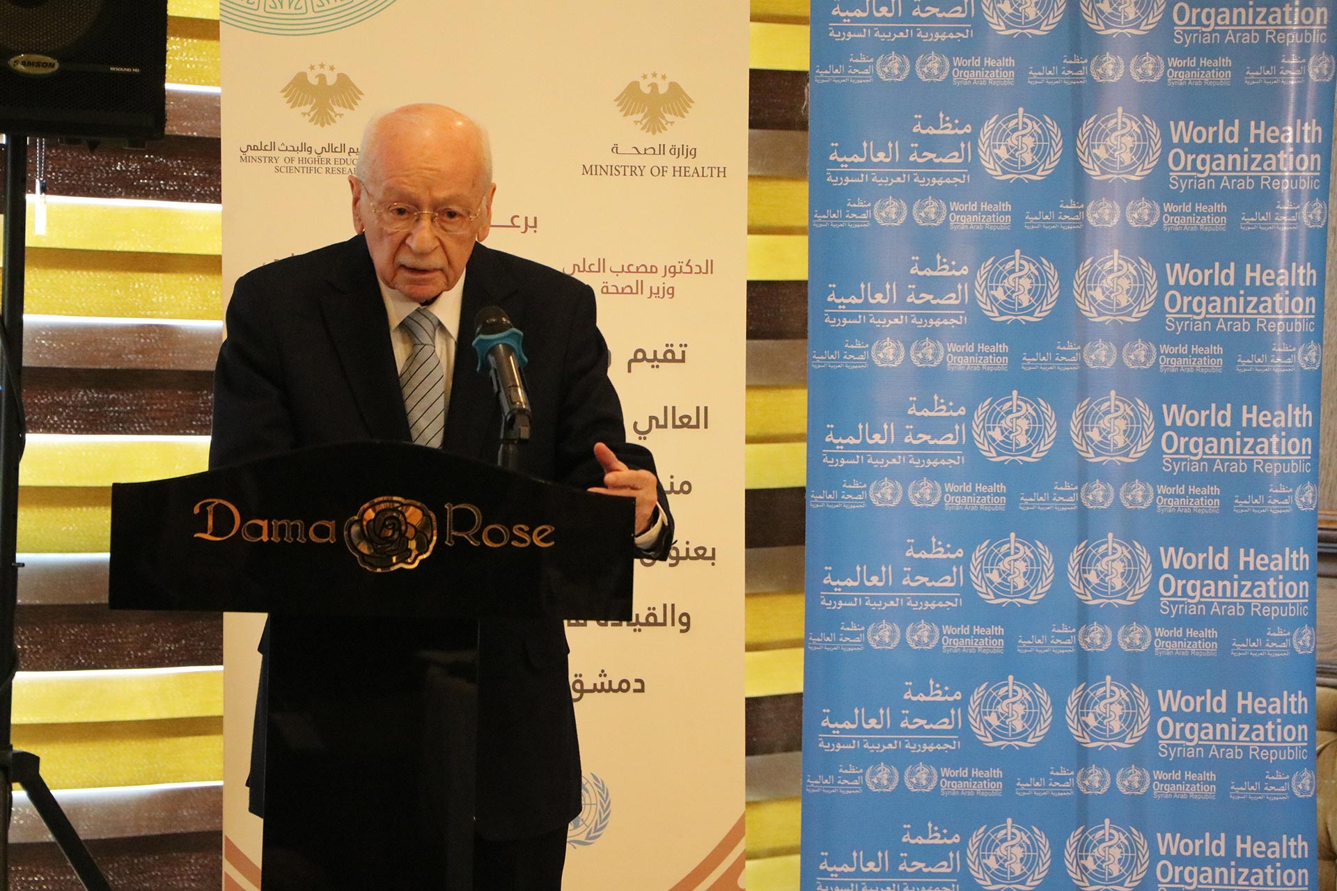 A speaker standing at a podium labeled “Dama Rose” delivers remarks during an event, with Ministry of Health and WHO banners displayed behind them.