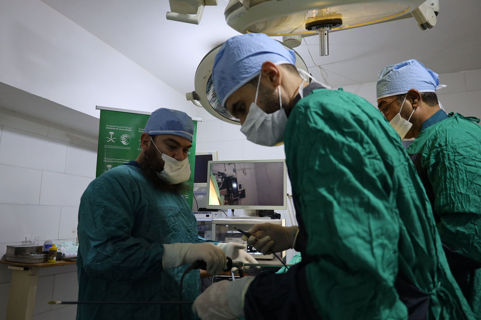 Surgeons in green gowns and blue surgical caps perform a procedure using specialized laparoscopic equipment, with monitors and overhead surgical lights visible in the operating room.